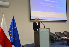 Przy mównicy stoi kobieta i przemawia.